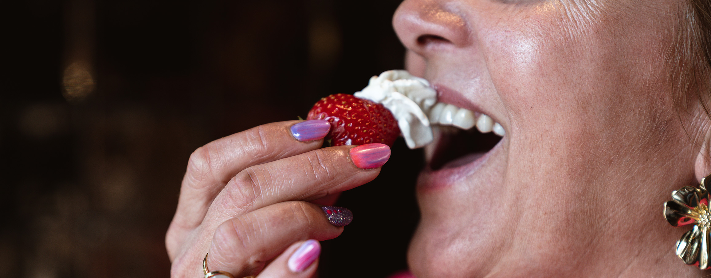 Vrouw met roze nagels eet lachend een aardbei met slagroom.